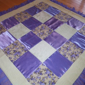 May include: A patchwork quilt with a cream background and purple satin squares. The quilt has a floral pattern in the center of each square. The quilt is edged with a light blue ruffle.