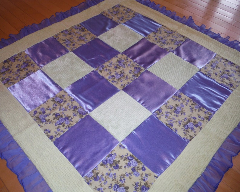 May include: A patchwork quilt with a cream background and purple satin squares. The quilt has a floral pattern in the center of each square. The quilt is edged with a light blue ruffle.