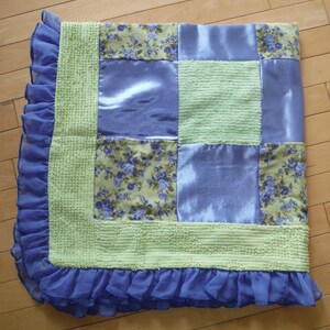 May include: A green and purple patchwork quilt with a ruffled edge. The quilt is made of different fabrics, including satin, floral print, and textured fabric.