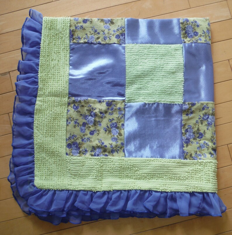 May include: A green and purple patchwork quilt with a ruffled edge. The quilt is made of different fabrics, including satin, floral print, and textured fabric.