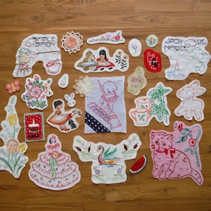 May include: A collection of vintage embroidered patches and appliques, featuring various designs such as floral patterns, children, animals, and text. The color palette includes white, pink, red, and green, with some patches having lace or other decorative elements.