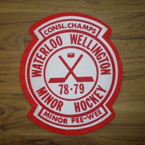 May include: Red and white circular patch with the text "Waterloo Wellington Consl. Champs 78-79 Minor Hockey Minor Pee-Wee" and a crossed hockey stick graphic.