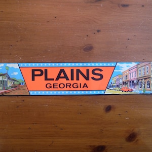 May include: A blue and orange bumper sticker with the text "PLAINS GEORGIA" and images of a building and a street scene. The sticker says "BUMPER STICKER" on the right side.