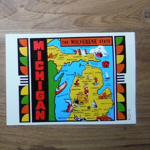 May include: A vintage postcard featuring a colorful map of Michigan, labeled "The Wolverine State". The map highlights major cities and landmarks, including Detroit, Grand Rapids, and Mackinac Island. The postcard has a red, white, and blue border with the word "MICHIGAN" in large black letters.