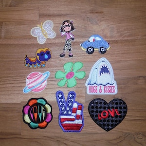 Könnte beinhalten: Eine Sammlung von gestickten Aufnähern in verschiedenen Formen und Designs. Enthält einen Schmetterling, ein Mädchen, ein blaues Auto, eine Zeichentrickfigur, einen Planeten, eine Blume, einen Hai mit "Hugs & Kisses", eine Blume mit "LOVE", ein Friedenszeichen und ein Herz mit "LOVE".