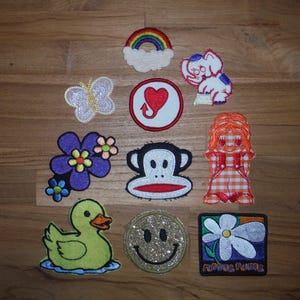 May include: A collection of embroidered patches featuring various designs. Includes a rainbow, butterfly, heart, elephant, flower, monkey, doll, duck, smiley face, and a flower with text. Patches are in various colors and shapes.