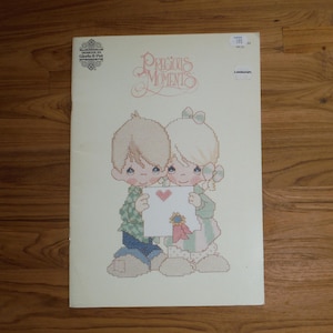 Puede incluir: Un folleto de patrones de punto de cruz Precious Moments con un diseño de dos niños sosteniendo una tarjeta blanca con un corazón rojo y una cinta azul y roja. La portada incluye el texto Precious Moments y Designs by Gloria & Pat.