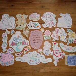 May include: A selection of embroidered patches in diverse shapes, such as a swan, flowers, and figures. The patches feature pastel colours and delicate stitching, with some including text. The patches are arranged on a wooden surface.