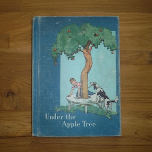 May include: A blue hardback book titled Under the Apple Tree, featuring an illustration of a child and a black and white dog sitting on a circular bench built around the trunk of an apple tree with small red apples in the green foliage.