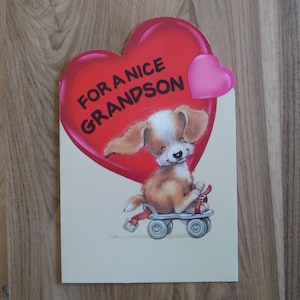 Pode incluir: Um cartão vermelho em forma de coração com o texto "For a Nice Grandson" e um pequeno coração rosa no canto superior direito. Um cachorrinho de desenho animado usando uma coleira vermelha e andando de patins está no centro do cartão.