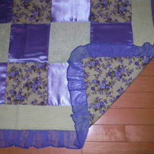 May include: A patchwork quilt with a purple and cream color scheme. The quilt is made of different fabrics, including satin, floral print, and a textured fabric. The quilt has a blue ruffle trim around the edges.