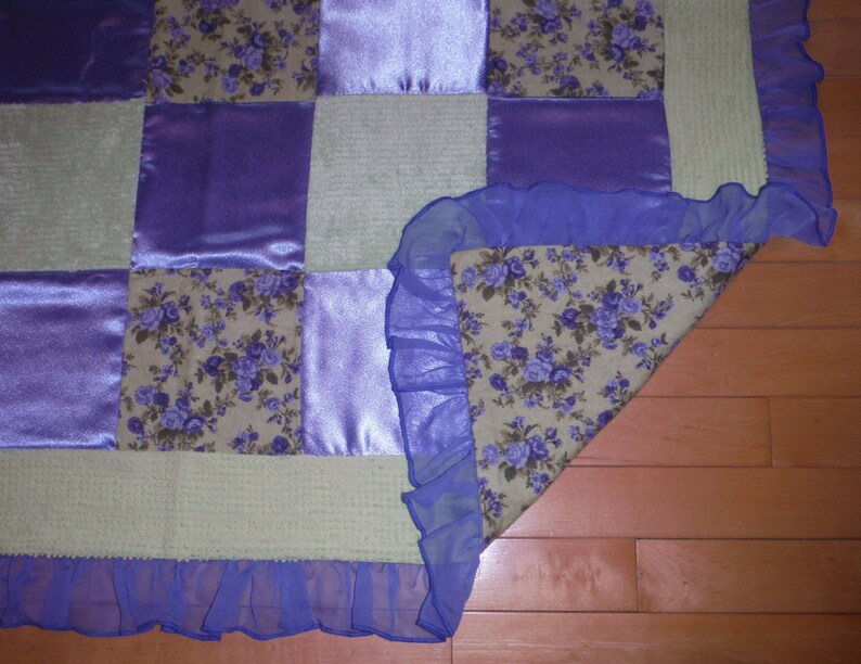 May include: A patchwork quilt with a purple and cream color scheme. The quilt is made of different fabrics, including satin, floral print, and a textured fabric. The quilt has a blue ruffle trim around the edges.