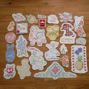 May include: A selection of vintage embroidered patches and appliques, showcasing diverse designs such as flowers, animals, and text. The colour palette includes white, red, blue, and green, with detailed stitching. The patches are displayed on a wooden surface.