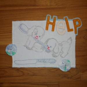 May include: Embroidered artwork featuring two cartoon puppies with blue and pink accents, a knife, and the word "HELP" in orange and white. Two small, round, fabric embellishments are also included.