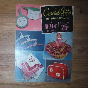 May include: A vintage magazine cover titled "Crochet Gifts and Bazaar Novelties" featuring various crochet projects. The cover includes illustrations of a pan, hangers, a doll in a basket, a stack of handkerchiefs, and a red toaster cover with daisy accents.