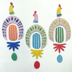 Peut inclure: Trois mobiles suspendus colorés en forme de poulets. Chaque mobile présente un poulet jaune, rose ou gris avec une crête rouge et un bec bleu. Les mobiles sont décorés de formes géométriques et de motifs de différentes couleurs, notamment le vert, le bleu, l'orange et le jaune.