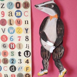 Puede incluir: Una figura articulada de tej&oacute;n con pajarita naranja y zapatos amarillos sobre fondo rosa. Junto a ella, un p&oacute;ster con letras, n&uacute;meros y s&iacute;mbolos en c&iacute;rculos de colores.
