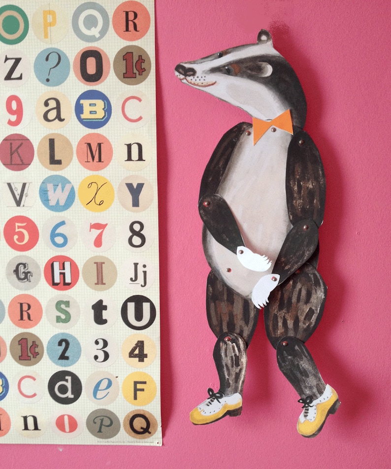 Puede incluir: Una figura articulada de tej&oacute;n con pajarita naranja y zapatos amarillos sobre fondo rosa. Junto a ella, un p&oacute;ster con letras, n&uacute;meros y s&iacute;mbolos en c&iacute;rculos de colores.