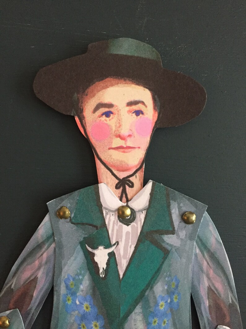 May include: A paper doll cutout of a person wearing a wide-brimmed black hat, a green jacket with gold buttons, a white shirt with a black bow tie, and a skull pin. The jacket is decorated with blue forget-me-not flowers.