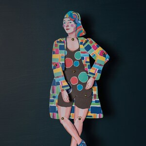 May include: A paper doll wearing a colourful geometric patterned coat and a black and white polka dot swimsuit. The doll has a blue headscarf and is standing with its legs crossed.