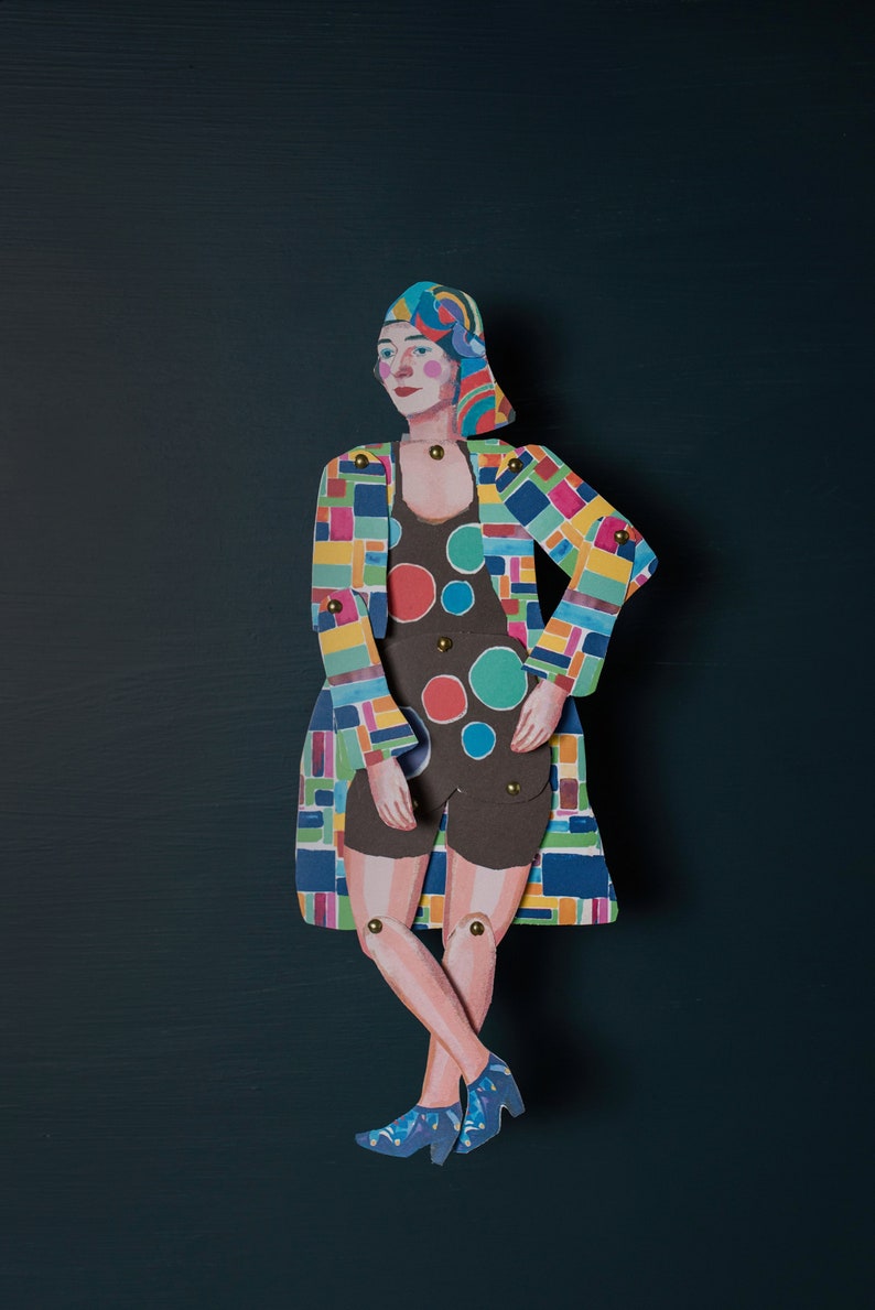 May include: A paper doll wearing a colourful geometric patterned coat and a black and white polka dot swimsuit. The doll has a blue headscarf and is standing with its legs crossed.