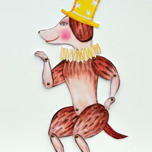 May include: A whimsical, hand-painted dog puppet with articulated joints. The dog wears a yellow top hat adorned with white stars and a ruffled collar. Its body is primarily brown with pink cheeks, and the design is reminiscent of a vintage toy.
