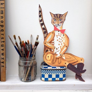 May include: A hand-painted cardboard cat puppet with a red bow tie, sitting on a blue and white checked box. The cat is brown and tan with black spots and has blue eyes. There are paintbrushes in a glass jar behind the cat.