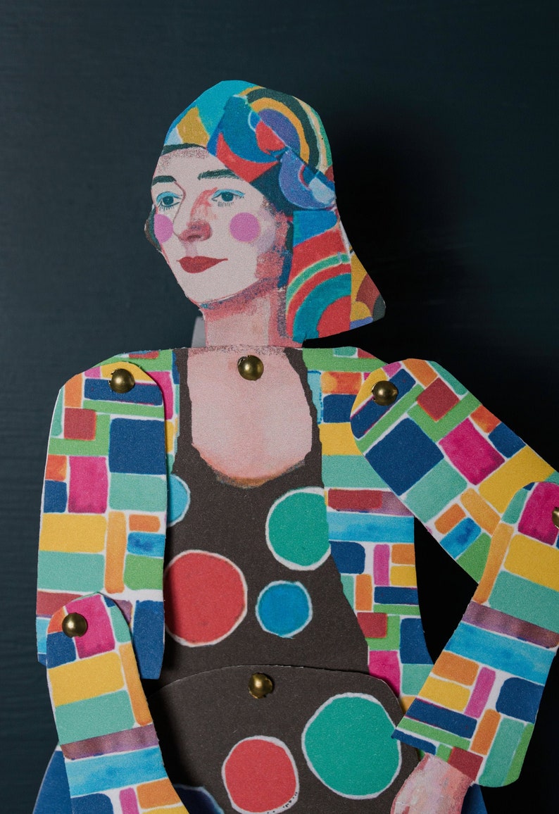 May include: A colourful paper doll with a geometric patterned dress and a colourful head scarf. The doll has a painted face with pink cheeks and red lips.