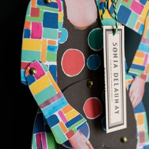 May include: A colourful paper doll with a geometric pattern and a tag that reads "SONIA DELAUNAY".
