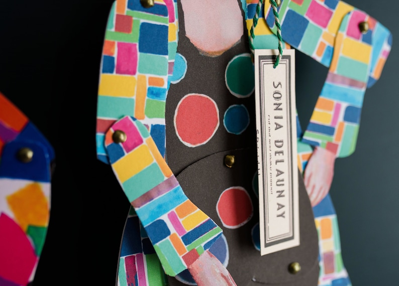 May include: A colourful paper doll with a geometric pattern and a tag that reads "SONIA DELAUNAY".