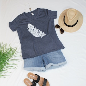 May include: A navy blue t-shirt with a white feather design. The shirt is paired with denim shorts, black sandals, a straw hat, and sunglasses.