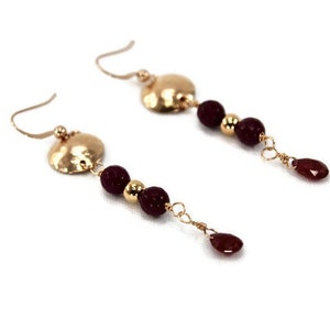 May include: Gold earrings with a hammered disc charm and burgundy beads. The earrings have a dangling burgundy gemstone at the bottom.