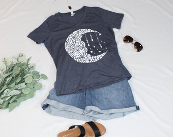 May include: A dark blue t-shirt with a white crescent moon design featuring a mandala pattern and hanging stars and raindrops. The shirt is paired with denim shorts and black sandals.