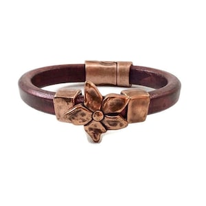 Könnte beinhalten: Ein braunes Lederarmband mit einem Kupferblumen-Anhänger. Das Armband hat einen Magnetverschluss.