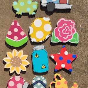 May include: A set of 12 wooden cutouts painted with various summer themes. The cutouts feature a watermelon, a lemon, a rose, a sunflower, a mason jar, a star, a flamingo, a pair of flip flops, a watering can, and a frog. Each cutout is decorated with white polka dots.