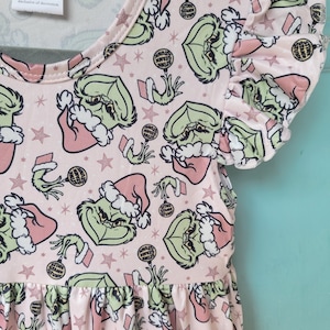 May include: A pink dress with a green Grinch print and the words "Stink Stank Stunk" repeated on the fabric. The dress has a ruffled neckline and short sleeves.