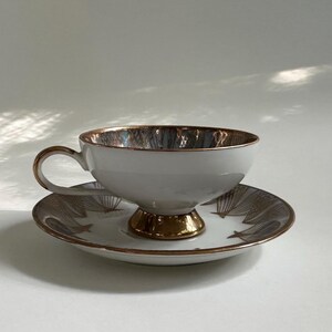 May include: A white porcelain teacup and saucer set featuring a metallic gold interior rim and a gold pedestal base. The saucer is decorated with a repeating pattern of thin, dark brown, fan-shaped lines radiating outward from the center.