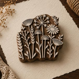 Könnte beinhalten: Ein brauner Holzstempel mit einem komplexen Schnitzdesign aus verschiedenen Wildblumen, Gräsern und einem kleinen Schmetterling. Die erhabene Oberfläche des Stempels zeigt detaillierte botanische Silhouetten, darunter Gänseblümchen.