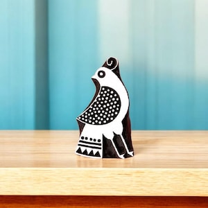 Puede incluir: Un sello de madera en blanco y negro con un diseño estilizado de un pájaro. El pájaro tiene un cuerpo blanco con lunares negros en su ala y un contorno negro. El sello está sobre una superficie de madera.