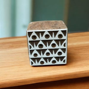 May include: A wooden block with a geometric pattern of triangles and circles. The block is painted with white and brown colors. The design is repeated across the surface, creating a symmetrical pattern. This block is likely used for printing or stamping.