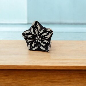 Könnte beinhalten: Ein Holzstempel mit einem schwarz-weißen Blumendesign. Der Stempel ist sternförmig mit einem detaillierten Blumenmuster. Er befindet sich auf einer hellbraunen Holzoberfläche mit hellblauem Hintergrund.
