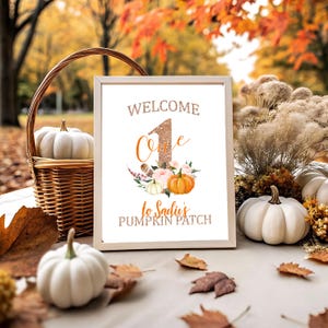 May include: A framed sign with the text "WELCOME One to Sadie's PUMPKIN PATCH" is surrounded by pumpkins, fall foliage, and a wicker basket. The sign features a glittery number one and floral accents, perfect for a fall-themed celebration.