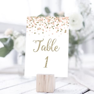 May include: White table number card with gold glitter text that says "Table 1" and a pink and gold confetti design. The card is on a wooden stand.