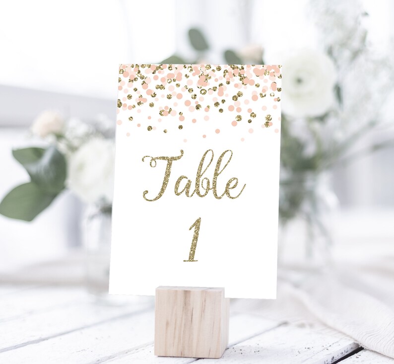 May include: White table number card with gold glitter text that says "Table 1" and a pink and gold confetti design. The card is on a wooden stand.