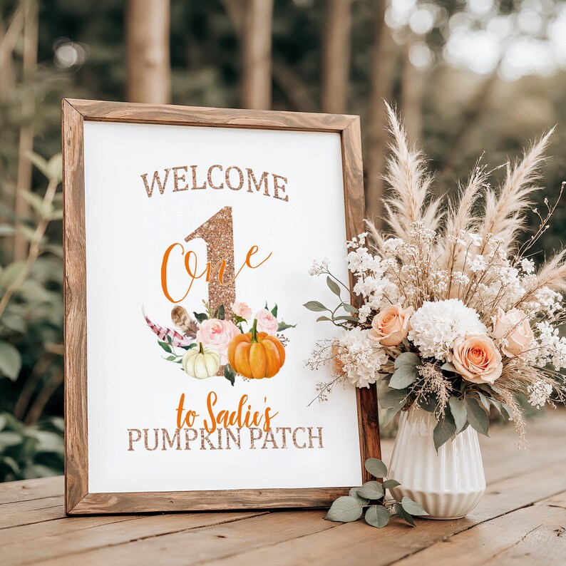 May include: A framed sign with the words "WELCOME One to Sadie's PUMPKIN PATCH" in glittery gold lettering. The sign features a large number one, pumpkins, and floral accents. A vase of flowers sits beside the sign.