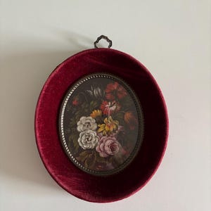 May include: Oval framed floral art. The artwork features a bouquet of white, pink, yellow, and red flowers. The frame is a deep red velvet with a dark metal decorative trim and hanging loop.
