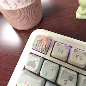 May include: A white keyboard with custom keycaps. One keycap features a gray knitted design with a pink bow. Other keycaps have light blue cat-themed designs. A pink and white floral patterned tumbler is in the background.