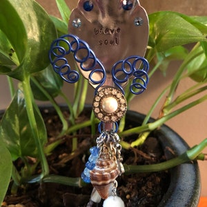 May include: A silver spoon with blue wire embellishments and rhinestones, a seashell, and beads. The spoon has the words "brave soul" stamped on it.