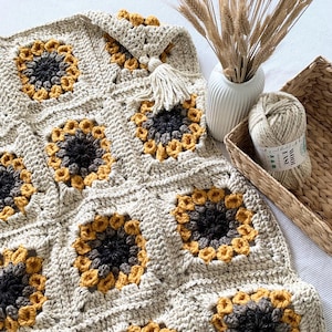May include: A crocheted blanket with a sunflower pattern in shades of beige, yellow, and gray. The blanket is made with a chunky yarn and has a tassel on one corner.