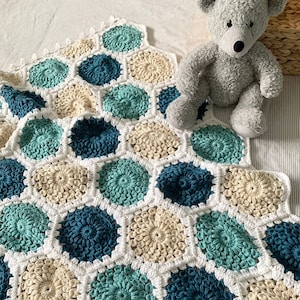 Puede incluir: Una manta de bebé tejida a crochet con un patrón hexagonal en tonos de azul verdoso, azul y crema.